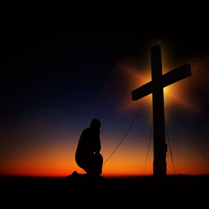 A person kneeling in prayer before a cross at sunset